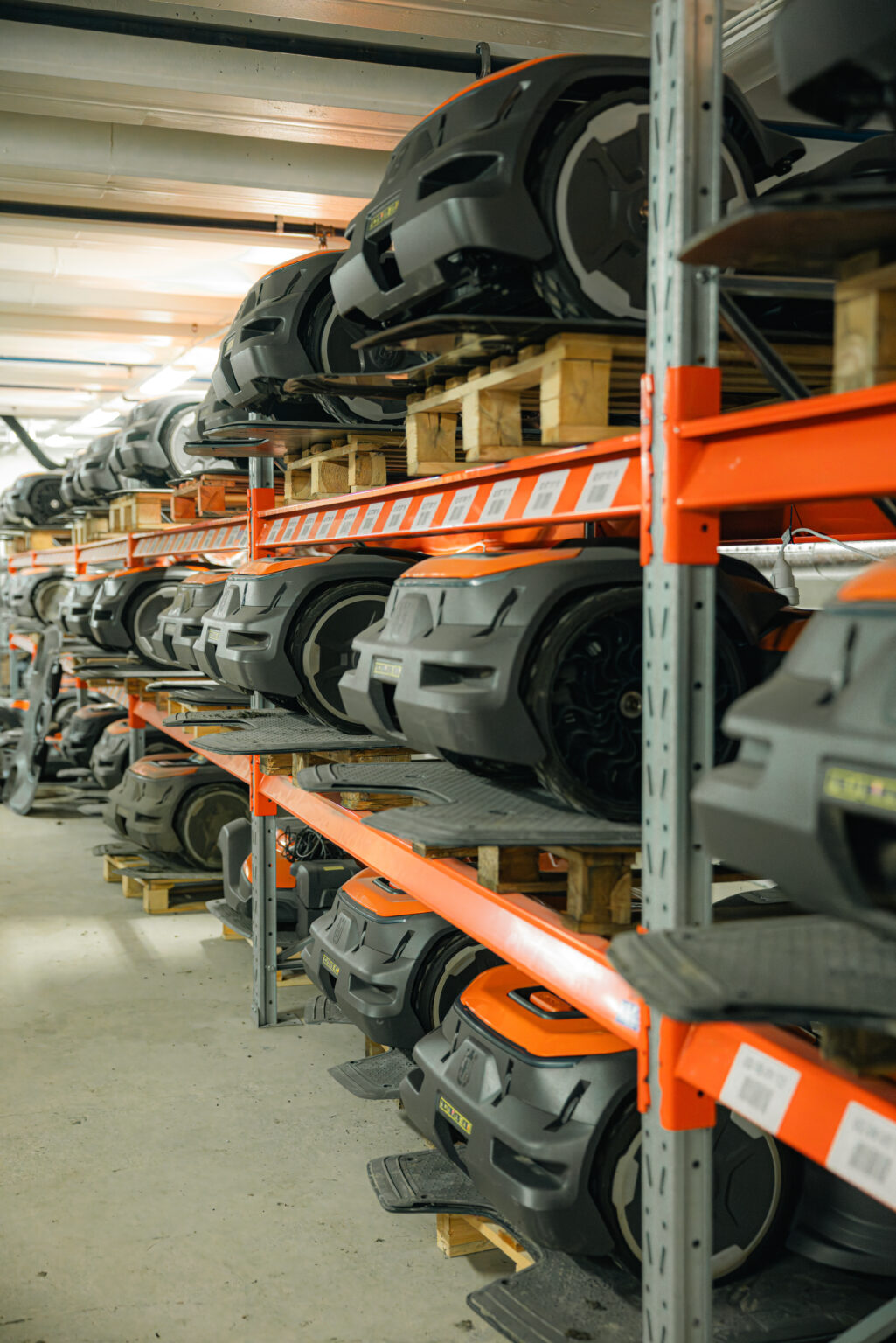 Robotgräsklippare från Husqvarna och andra märken lagrade på orange pallställ i verkstadslokal