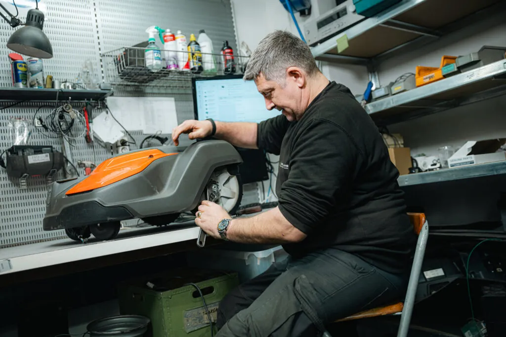Tekniker reparerar robotgräsklippare med orange hölje i välutrustad verkstad med verktyg och reservdelar